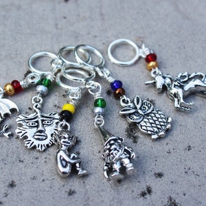 May include: Six silver stitch markers with colorful beads and charms. The charms include a dragon, a sun face, a gnome, a monkey, an owl, and a unicorn.