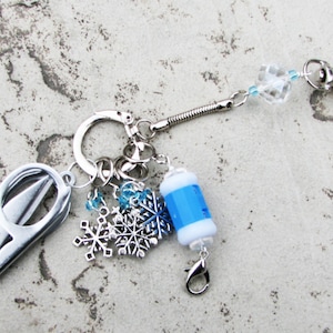 May include: A silver key chain with a variety of charms, including a pair of small scissors, a blue and white bead, and several snowflake charms. The key chain also features a clear crystal bead and a silver spring ring.