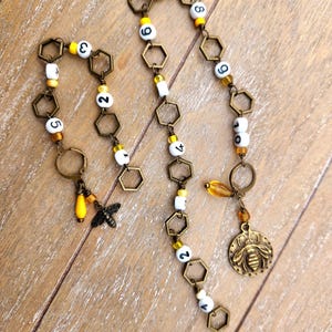 May include: Handmade bee-themed jewelry featuring bronze-colored hexagonal links, white beads with black numbers, and yellow accents. Includes a bee charm, a bee pendant, and a teardrop-shaped bead. The jewelry is displayed on a wooden surface.