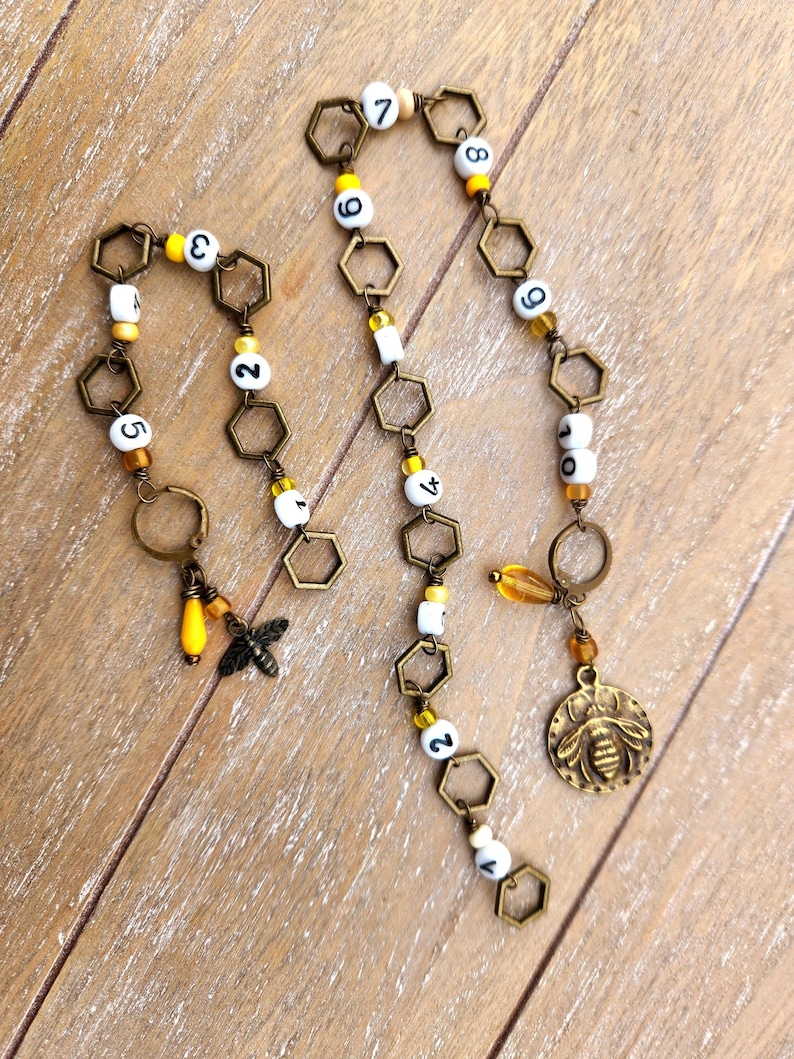 May include: Handmade bee-themed jewelry featuring bronze-colored hexagonal links, white beads with black numbers, and yellow accents. Includes a bee charm, a bee pendant, and a teardrop-shaped bead. The jewelry is displayed on a wooden surface.