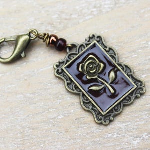 May include: An antique bronze-toned charm with a rectangular, burgundy-colored center featuring a rose design. The charm has an ornate frame and is attached to a lobster clasp and a string of dark red beads.