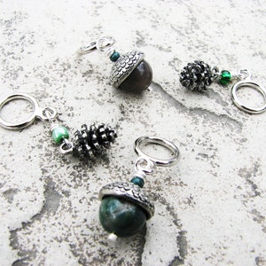 May include: Three silver acorn and pine cone stitch markers with green beads. The acorn stitch markers have a silver cap and a green stone in the center. The pine cone stitch marker has a silver pine cone with a green bead.