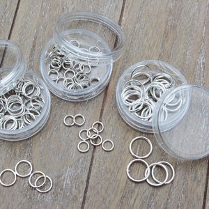 May include: Three clear plastic containers filled with silver jump rings. The containers are on a wooden surface. There are also some jump rings scattered on the surface.