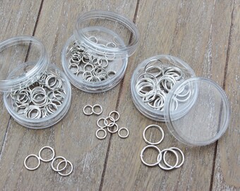 May include: Three clear plastic containers filled with silver jump rings. The containers are on a wooden surface. There are also some jump rings scattered on the surface.