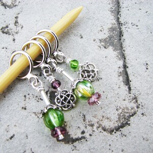 May include: Four silver stitch markers with green and purple glass beads. The markers are attached to a wooden knitting needle.