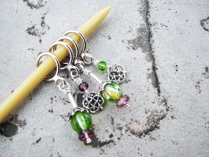 May include: Four silver stitch markers with green and purple glass beads. The markers are attached to a wooden knitting needle.