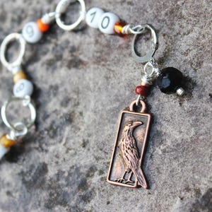 May include: A stitch marker set featuring silver-toned rings and white beads printed with black numbers. The end of the chain includes a copper-colored rectangular charm depicting a raven, accented by a small red bead and a faceted black bead.