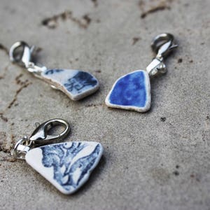May include: Three blue and white ceramic charms with silver clasps. The charms are shaped like a heart, a teardrop, and a rectangle. The charms are on a light gray background.