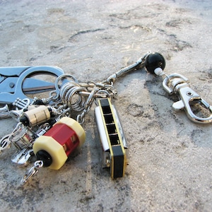 May include: A silver key chain with a variety of charms, including a small harmonica, a pair of scissors, and a small musical instrument. The key chain also has a black bead and a silver clasp.