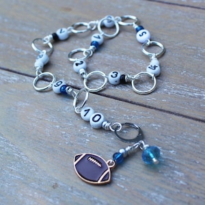 May include: A silver chain bracelet with white beads featuring black numbers and blue accents. A football charm with a brown and gold outline hangs from the bracelet, along with a blue bead.