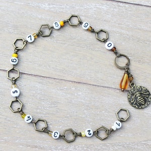 May include: A bronze-toned necklace featuring a series of hexagonal links with white beads displaying numbers. A charm with a bee design and a teardrop-shaped amber bead hangs from the necklace. The necklace is on a light wood surface.