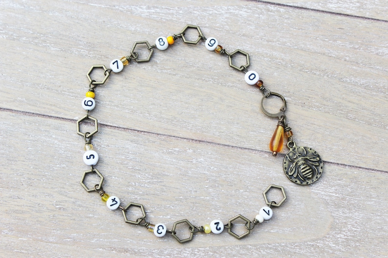 May include: A bronze-toned necklace featuring a series of hexagonal links with white beads displaying numbers. A charm with a bee design and a teardrop-shaped amber bead hangs from the necklace. The necklace is on a light wood surface.