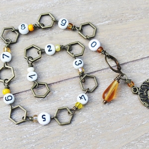 May include: A bronze-toned bracelet featuring hexagonal links and white beads with black numbers. A charm with a bee design and a teardrop-shaped bead dangle from the bracelet. The bracelet is on a wooden surface.