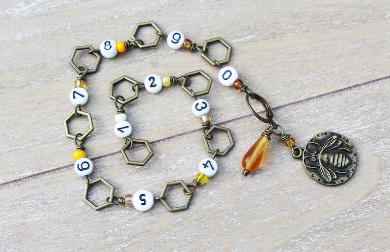 May include: A bronze-toned bracelet featuring hexagonal links and white beads with black numbers. A charm with a bee design and a teardrop-shaped bead dangle from the bracelet. The bracelet is on a wooden surface.