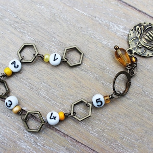 May include: A handmade bracelet featuring a bee charm and hexagonal links. The bracelet includes white beads with black numbers 1 through 5, interspersed with yellow and orange beads. The charm is a bronze-colored disc with a bee design.