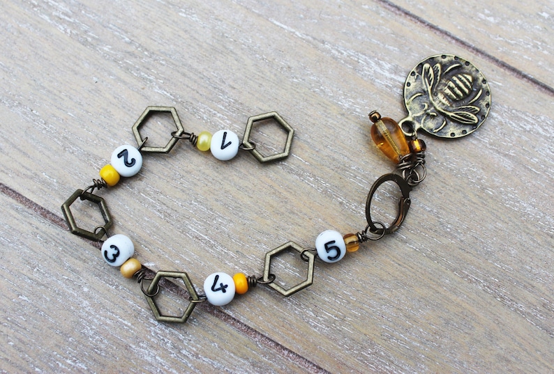 May include: A handmade bracelet featuring a bee charm and hexagonal links. The bracelet includes white beads with black numbers 1 through 5, interspersed with yellow and orange beads. The charm is a bronze-colored disc with a bee design.