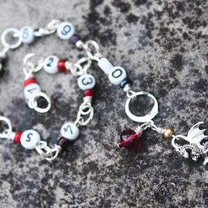 May include: A silver-toned bracelet with white beads featuring black numbers and red accents. A dragon charm and a red crystal dangle from the bracelet. The bracelet is on a textured gray surface.
