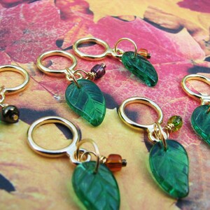 May include: Six gold stitch markers with green glass leaf charms. The charms have a textured surface and are attached to the markers with gold wire. The markers are on a background of fall leaves.