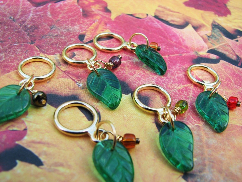May include: Six gold stitch markers with green glass leaf charms. The charms have a textured surface and are attached to the markers with gold wire. The markers are on a background of fall leaves.