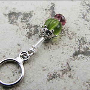 May include: A silver charm with a green and pink glass bead. The bead has a silver cap with decorative details. The charm is attached to a silver ring.