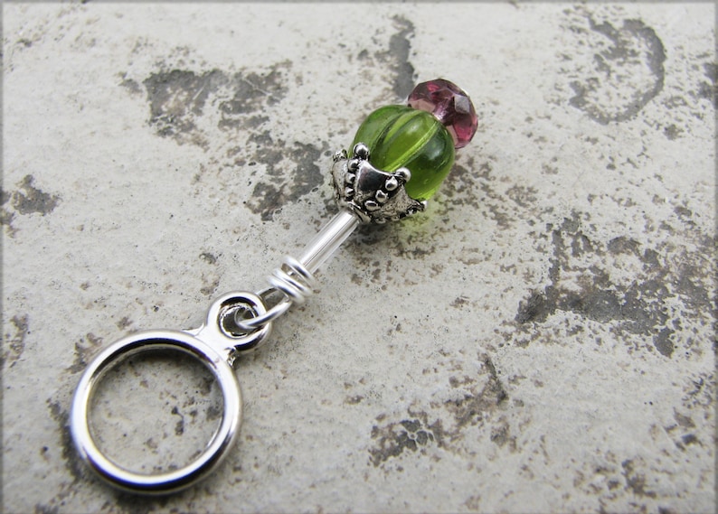 May include: A silver charm with a green and pink glass bead. The bead has a silver cap with decorative details. The charm is attached to a silver ring.