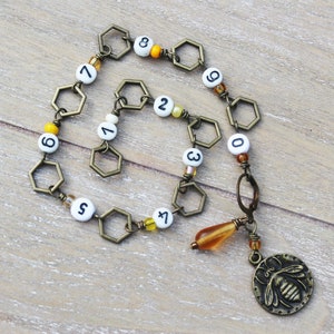 May include: A handmade necklace featuring a series of bronze-colored hexagonal links interspersed with white beads displaying numbers. The necklace includes a charm with a bee design and a teardrop-shaped amber bead.