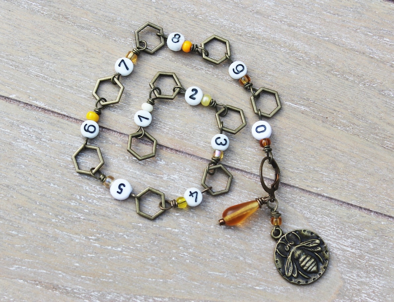 May include: A handmade necklace featuring a series of bronze-colored hexagonal links interspersed with white beads displaying numbers. The necklace includes a charm with a bee design and a teardrop-shaped amber bead.