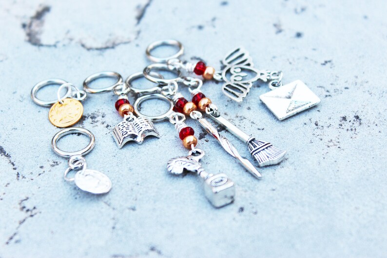Peut inclure: Ensemble de six marqueurs de points de tricot en argent avec des breloques, dont un livre, un balai, un hibou, une lettre, une fiole de potion et une pi&egrave;ce d'or. Les breloques sont attach&eacute;es aux marqueurs avec des perles rouges et dor&eacute;es.