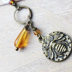 May include: A bronze-toned earring featuring a round charm with a bee design. The earring includes a teardrop-shaped amber bead and small amber beads. The design has a vintage aesthetic.