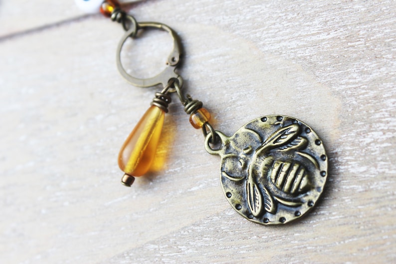 May include: A bronze-toned earring featuring a round charm with a bee design. The earring includes a teardrop-shaped amber bead and small amber beads. The design has a vintage aesthetic.
