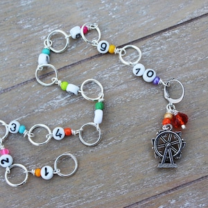 May include: A handmade beaded charm bracelet with silver rings and white beads with numbers 1 through 10. The bracelet has colorful beads and a Ferris wheel charm. The bracelet is on a wooden surface.