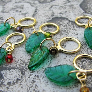 May include: Six green glass leaf stitch markers with gold rings and colorful beads. The leaves have a raised vein pattern.