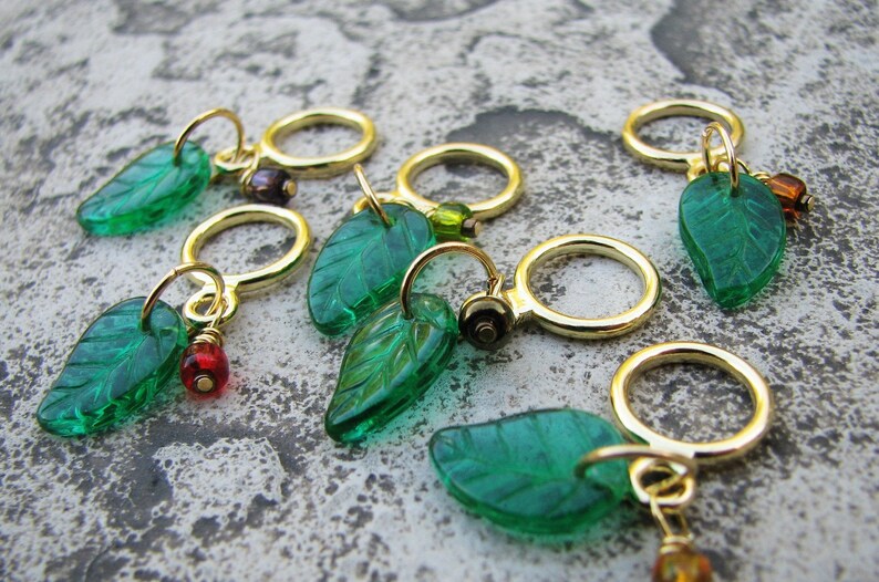 May include: Six green glass leaf stitch markers with gold rings and colorful beads. The leaves have a raised vein pattern.
