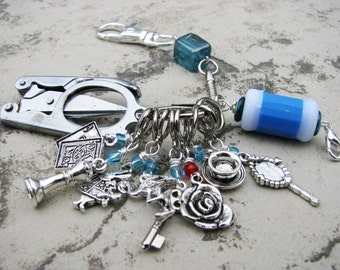 May include: A silver charm bracelet with a variety of charms, including a playing card, a teacup, a rose, a key, and a mirror. The bracelet also features a blue and white striped charm and a silver multi-tool.