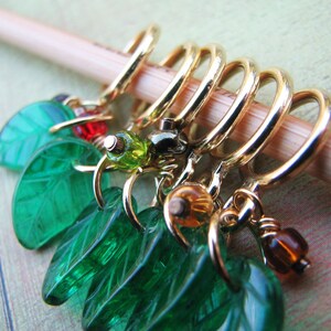 May include: A set of six gold stitch markers with green leaf charms. The charms are made of glass and have a textured surface. The stitch markers are attached to the charms with wire.