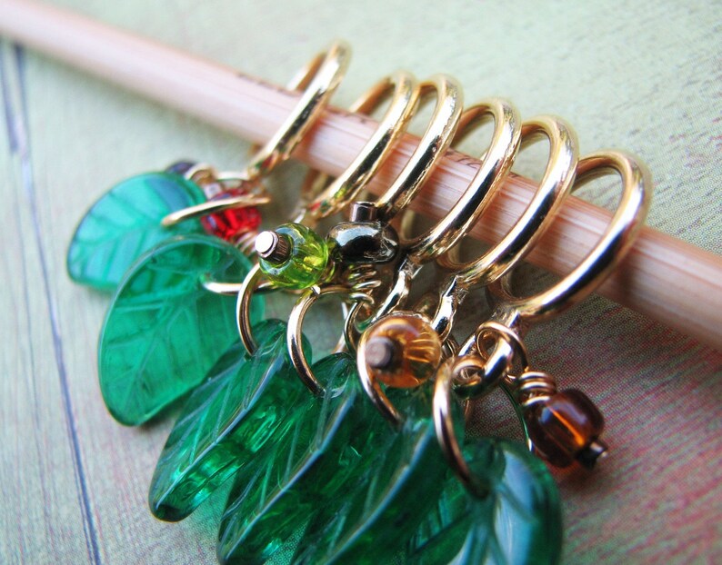 May include: A set of six gold stitch markers with green leaf charms. The charms are made of glass and have a textured surface. The stitch markers are attached to the charms with wire.