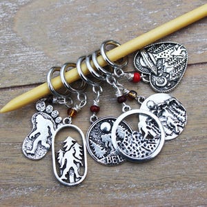 May include: Set of six silver knitting stitch markers with various nature-themed designs, including a footprint, a tree, a mountain range, and a heart with a tree inside. The markers are attached to a wooden knitting needle.