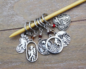 Puede incluir: Conjunto de seis marcadores de punto de tejido de plata con varios diseños de temática natural, que incluyen una huella, un árbol, una cordillera y un corazón con un árbol en su interior. Los marcadores están unidos a una aguja de tejer de madera.