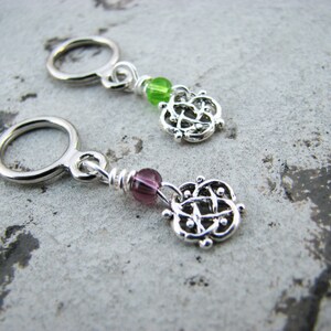 May include: Two silver stitch markers with a Celtic knot design. One marker has a green bead and the other has a purple bead.