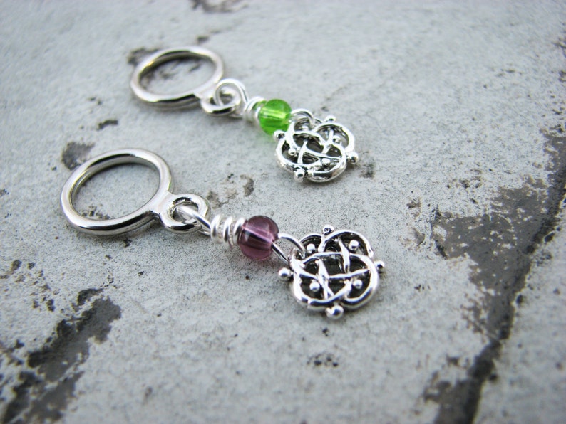 May include: Two silver stitch markers with a Celtic knot design. One marker has a green bead and the other has a purple bead.