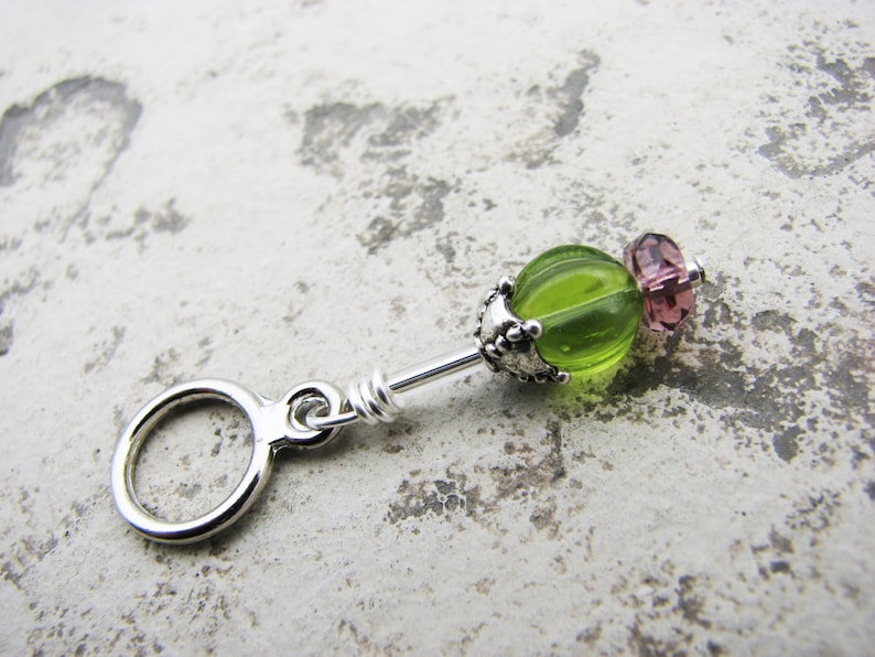 May include: A silver charm with a green glass bead and a pink glass bead. The charm has a ring at the top for attaching to a bracelet or necklace.