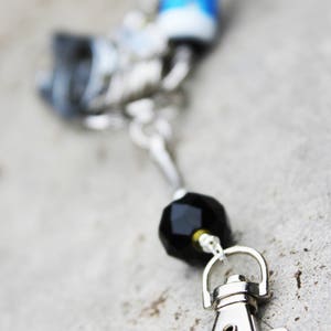 Peut inclure: Un porte-cl&eacute;s argent&eacute; avec une perle noire et un fermoir argent&eacute;. Le porte-cl&eacute;s a un charme bleu et blanc attach&eacute; &agrave; lui.