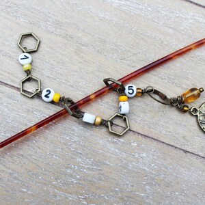 May include: A decorative hair stick with a dark amber finish. The stick features a chain of bronze-colored hexagonal charms, white beads with black numbers, and a bronze bee charm. The hair accessory is approximately 7 inches long.