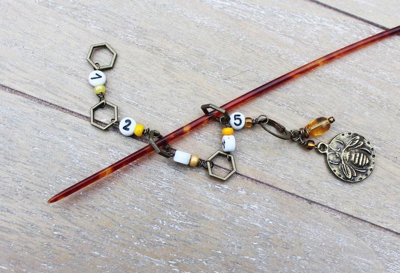 May include: A decorative hair stick with a dark amber finish. The stick features a chain of bronze-colored hexagonal charms, white beads with black numbers, and a bronze bee charm. The hair accessory is approximately 7 inches long.