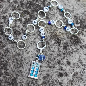 May include: A silver-toned charm bracelet featuring white beads with black numbers, connected by silver rings. A blue and silver TARDIS charm hangs from the bracelet, along with a blue bead. The bracelet is displayed on a textured gray surface.