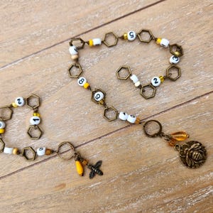 May include: A handmade bee-themed necklace and bracelet set. The necklace features a chain of hexagonal links, white and yellow beads with numbers, and a bee charm. The bracelet mirrors the necklace design.
