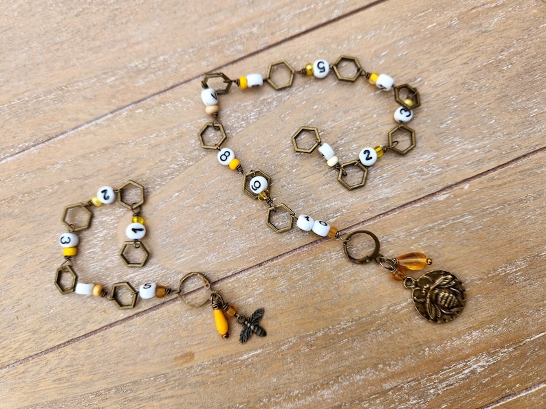 May include: A handmade bee-themed necklace and bracelet set. The necklace features a chain of hexagonal links, white and yellow beads with numbers, and a bee charm. The bracelet mirrors the necklace design.