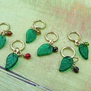 May include: Six green glass leaf stitch markers with gold rings and colorful beads. The markers are arranged on a green and brown wooden surface.