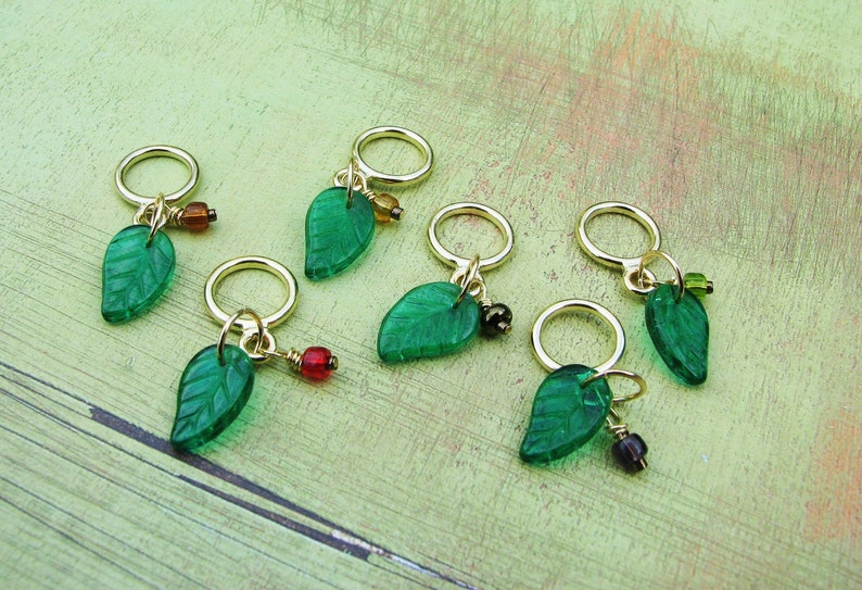 May include: Six green glass leaf stitch markers with gold rings and colorful beads. The markers are arranged on a green and brown wooden surface.