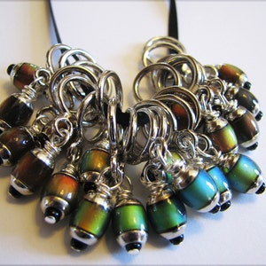 May include: A set of 14 iridescent stitch markers with silver rings and black beads. The markers are arranged in a cluster, with the rings and beads visible.